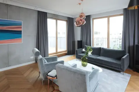 Nowoczesny salon z betonową ścianą, drewnianą podłogą w jodełkę, szarymi meblami i miedzianymi lampami. Lamele i beton tworzą industrialny klimat.