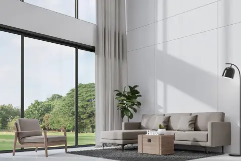Nowoczesny salon z wysokim sufitem, szara sofa, fotel i wielkie okna z widokiem na zieleń.