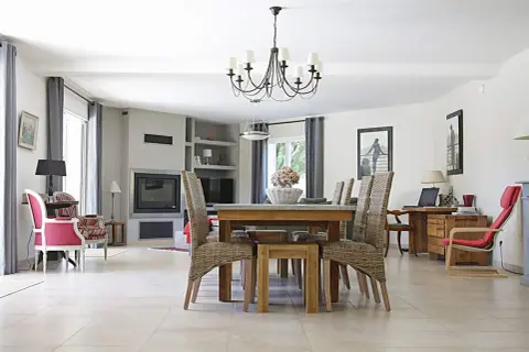 Elegancki, czarny żyrandol do salonu nad stołem z wiklinowymi krzesłami. W tle kominek i wygodne fotele.