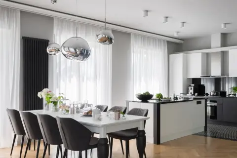 Elegancki styl glamour salon z kuchnią. Nowoczesne lampy, biała wyspa kuchenna i stół z szarymi krzesłami tworzą spójną całość.
