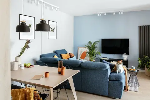 Przytulny, minimalistyczny salon w bloku. Niebieska kanapa, drewniany stół i kot na fotelu tworzą ciepłą atmosferę.